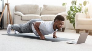 Un hombre haciendo flexiones en el salón de su casa mientras sigue una clase de entrenamiento online en una laptop