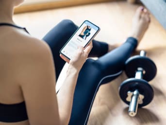 Mujer sentada en el suelo consultando una aplicación de entrenamiento personal en su móvil, con unas pesas a su lado.