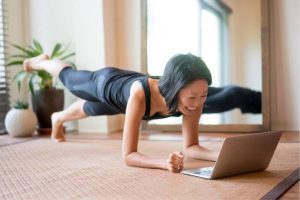 Mujer sonriendo mientras realiza una plancha con una pierna elevada, siguiendo su entrenamiento personal a través de un ordenador portátil.