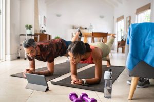 Pareja realizando una plancha en casa sobre colchonetas, siguiendo una sesión de entrenamiento online a través de una tablet