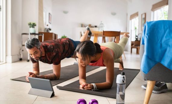 entrenamiento-hibrido-en-casa-pareja Pareja realizando una plancha en casa sobre colchonetas, siguiendo una sesión de entrenamiento online a través de una tablet