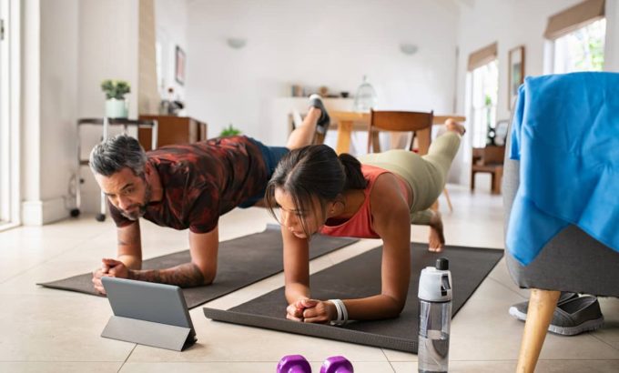 entrenamiento-hibrido-en-casa-pareja Pareja realizando una plancha en casa sobre colchonetas, siguiendo una sesión de entrenamiento online a través de una tablet
