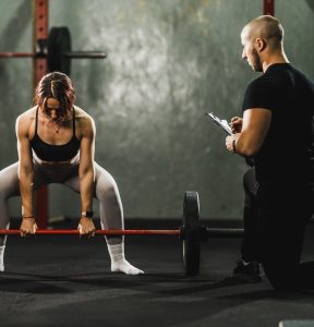 ntrenador personal masculino supervisando y corrigiendo la técnica de una mujer que realiza un peso muerto con barra en un gimnasio.