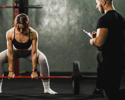 ntrenador personal masculino supervisando y corrigiendo la técnica de una mujer que realiza un peso muerto con barra en un gimnasio.