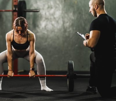 ntrenador personal masculino supervisando y corrigiendo la técnica de una mujer que realiza un peso muerto con barra en un gimnasio.