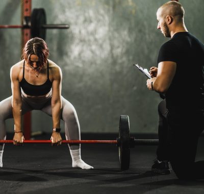 entrenador-personal-corrigiendo-peso-muerto. ntrenador personal masculino supervisando y corrigiendo la técnica de una mujer que realiza un peso muerto con barra en un gimnasio.