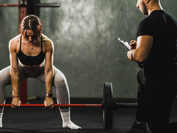 ntrenador personal masculino supervisando y corrigiendo la técnica de una mujer que realiza un peso muerto con barra en un gimnasio.